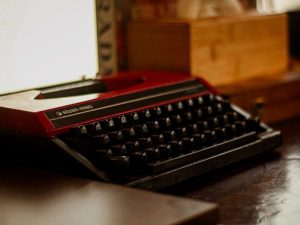 Red vintage typewriter resting on a dark wooden desk in a warmly lit interior.