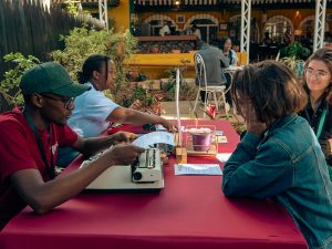 Live typewritten poetry session at an outdoor event with a poet using a typewriter while guests sit across the table listening and engaging shows why people turn to poetry during big life moments.